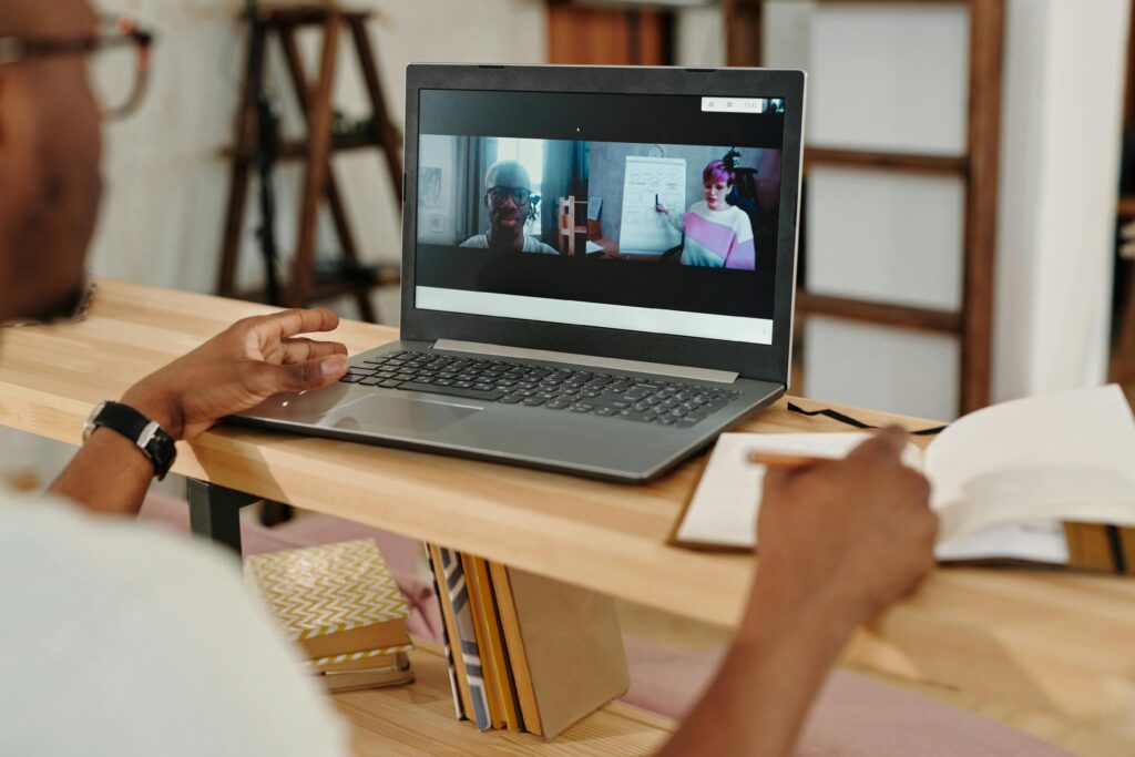 Adult using a laptop for video conferencing, taking notes during a remote meeting indoors.